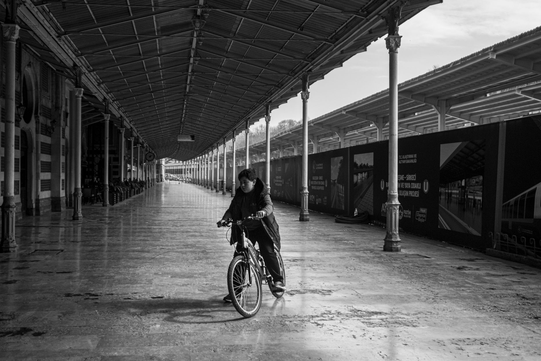 A person rides a bicycle in an empty Istanbul train station, in black and white.