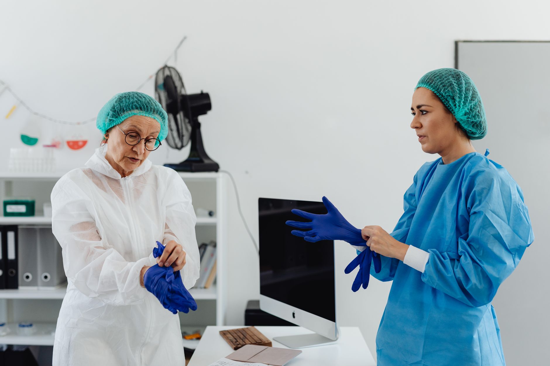 Two women healthcare workers in surgical attire putting on protective gloves in a clinic.