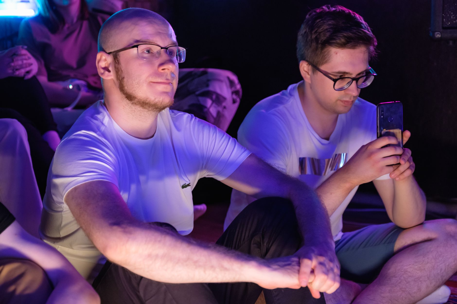 Two men seated indoors, one taking photos with a smartphone, amidst an engaged audience.