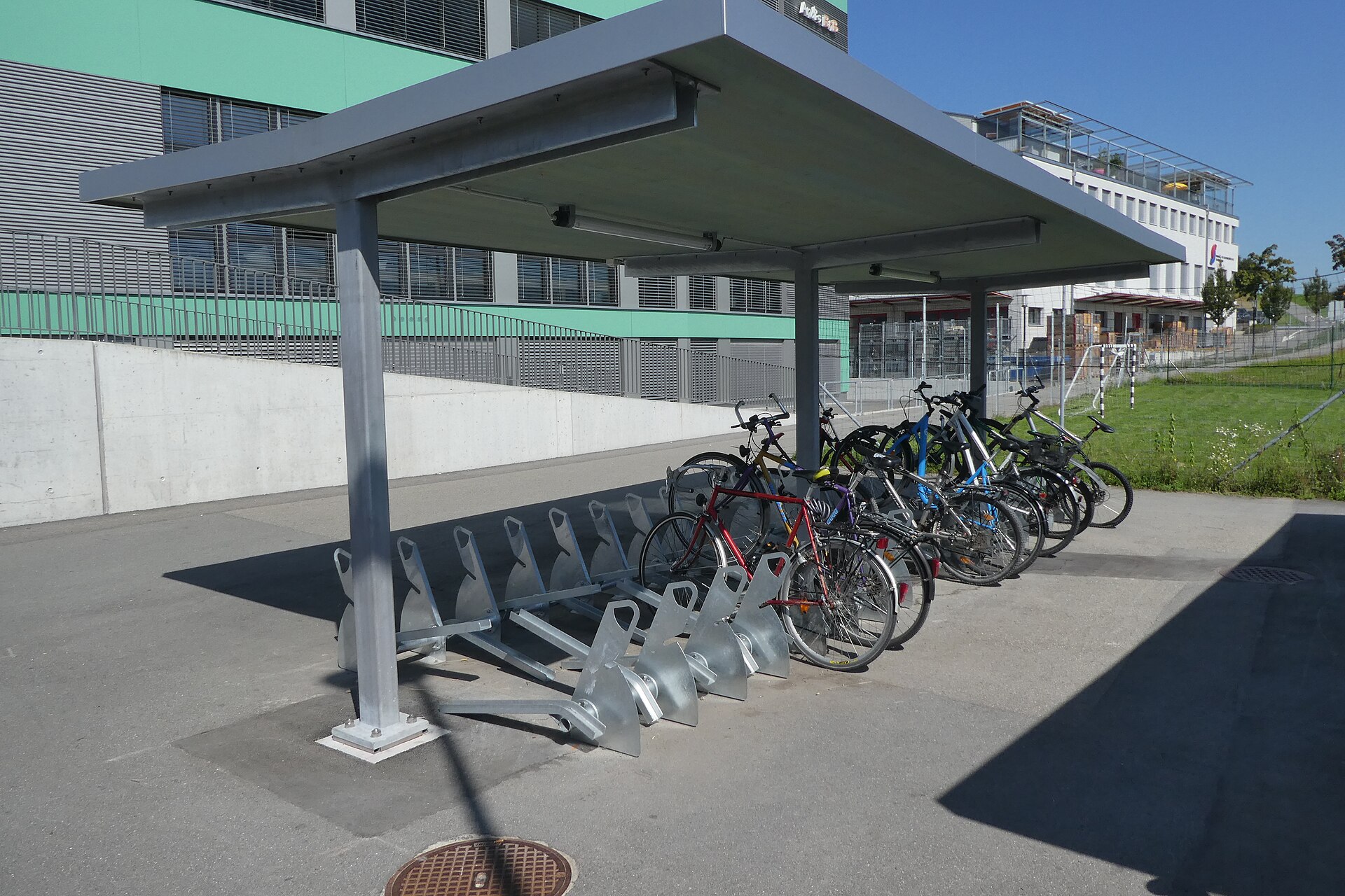 Abri à vélos couvert devant un bâtiment moderne, avec arceaux métalliques et plusieurs vélos stationnés sous un ciel dégagé.