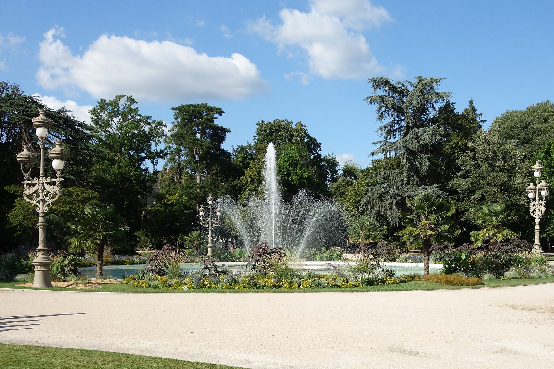 Jardin public arboré en été, avec une fontaine au centre d'un bassin, des massifs fleuris et des lampadaires anciens