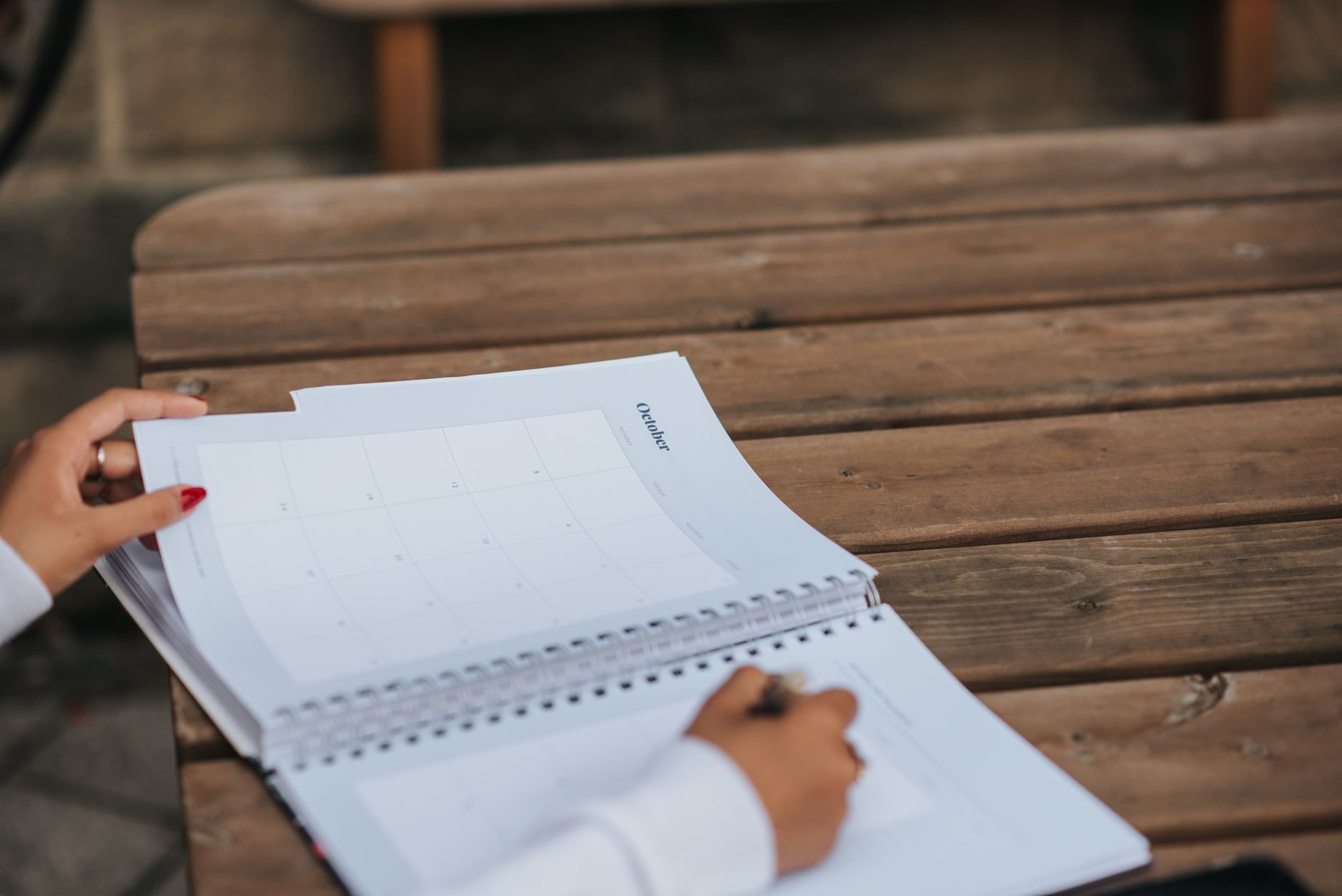 Mains écrivant dans un agenda à spirale ouvert sur le mois d’octobre, posé sur une table en bois en extérieur.