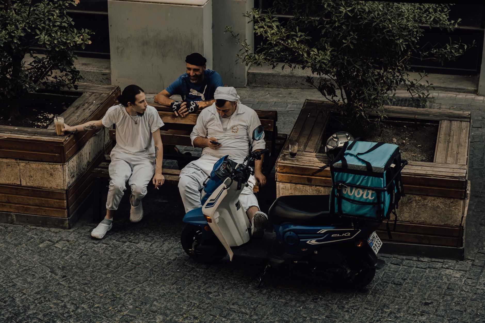 Trois hommes assis sur des bancs en bois dans une rue pavée, près d’un scooter bleu équipé d’un grand sac de livraison turquoise.